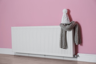 Modern radiator with knitted hat and scarf near color wall indoors. Central heating system