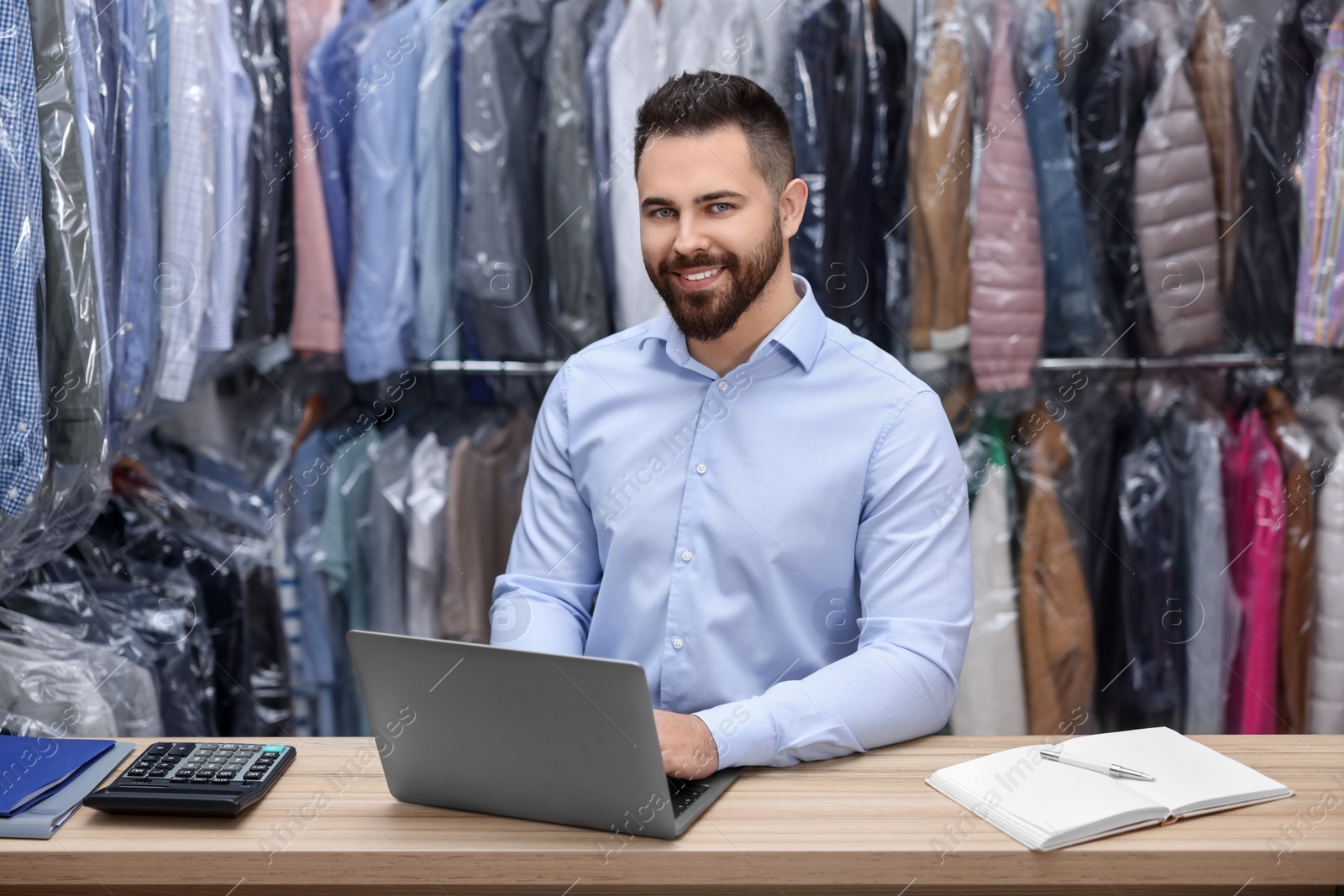 Photo of Dry-cleaning service. Happy worker using laptop at counter indoors