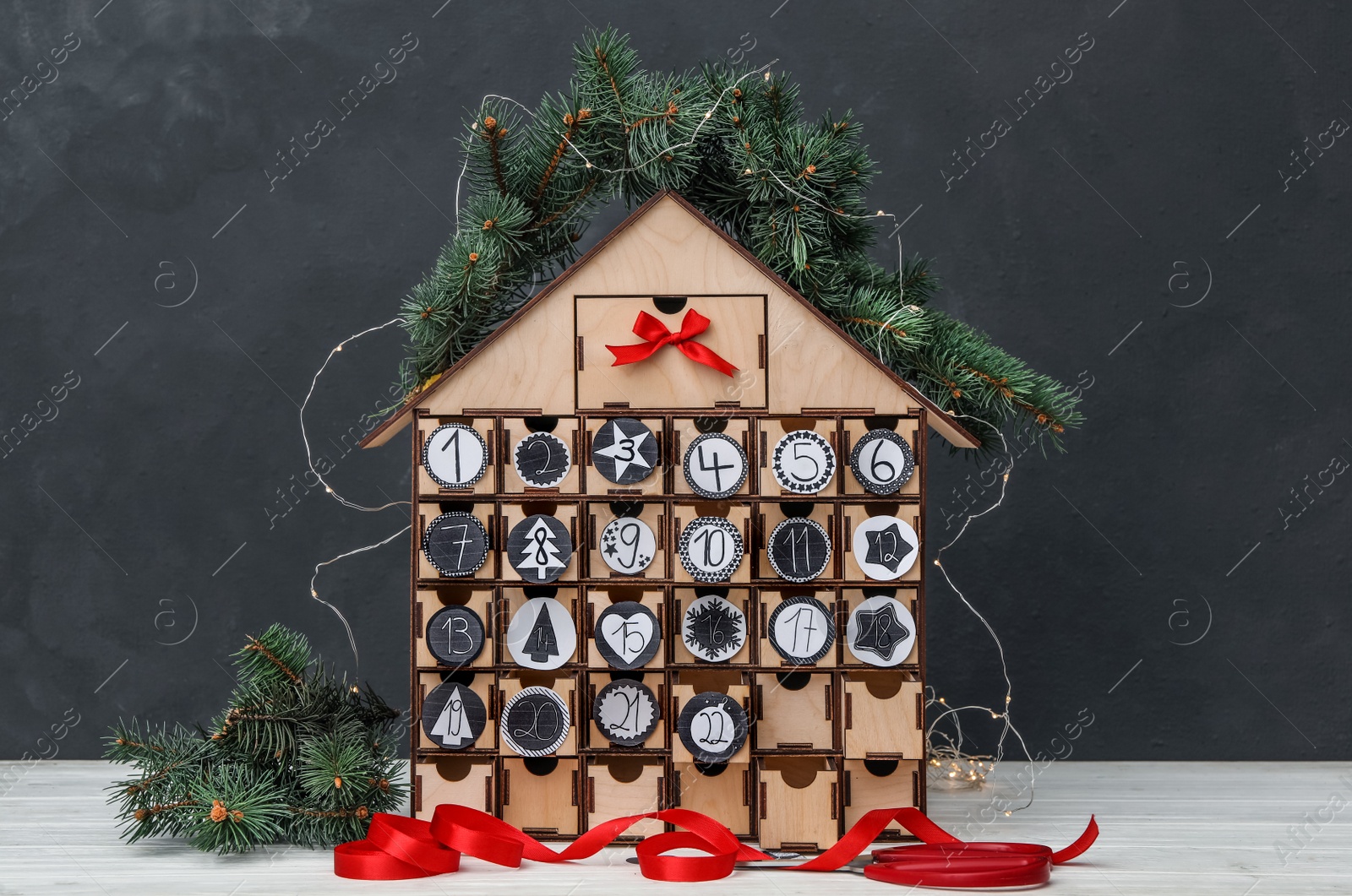 Photo of House shaped advent calendar with garland and fir tree on white wooden table against dark background