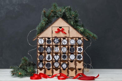 Photo of House shaped advent calendar with garland and fir tree on white wooden table against dark background