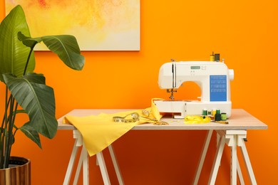 Photo of Modern sewing machine with cloth and craft accessories on wooden table near orange wall in room