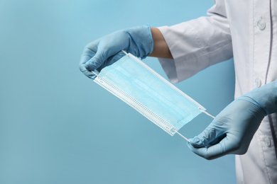 Photo of Doctor in latex gloves holding disposable face mask on light blue background, closeup. Protective measures during coronavirus quarantine