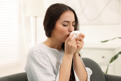 Photo of Sick woman with tissue blowing nose on sofa at home. Cold symptoms