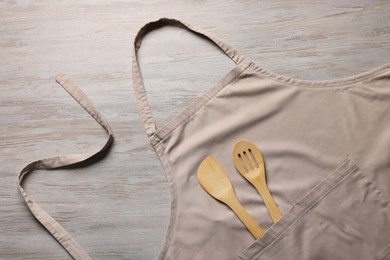 Stylish beige apron, spatula and slotted spoon on light wooden table, top view