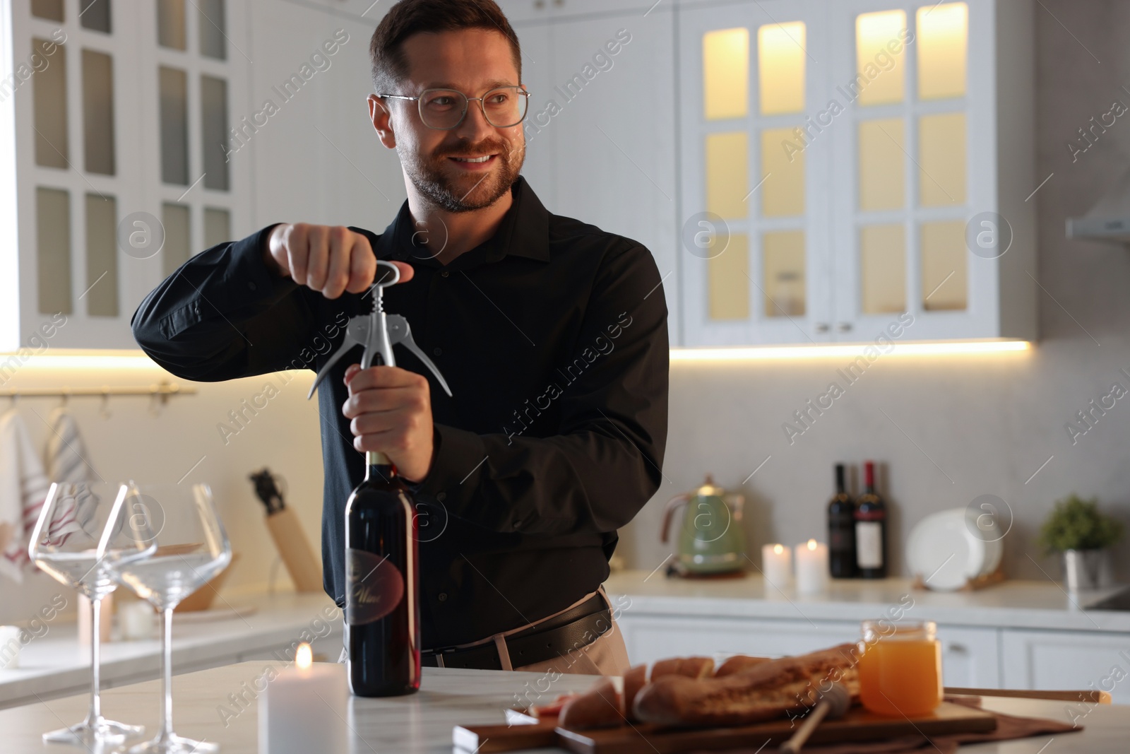 Photo of Romantic dinner. Man opening wine bottle with corkscrew at table in kitchen