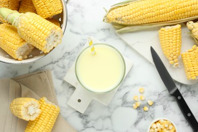 Photo of Tasty fresh corn milk in glass and cobs on white marble table, flat lay