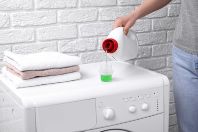 Photo of Woman pouring fabric softener from bottle into cap on washing machine near white brick wall, closeup