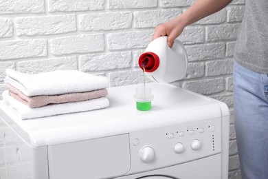 Woman pouring fabric softener from bottle into cap on washing machine near white brick wall, closeup