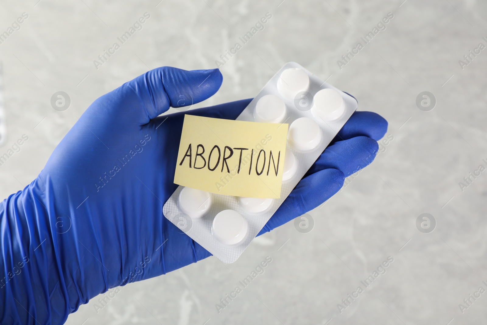 Photo of Doctor holding pills and paper card with word ABORTION on light grey background, closeup