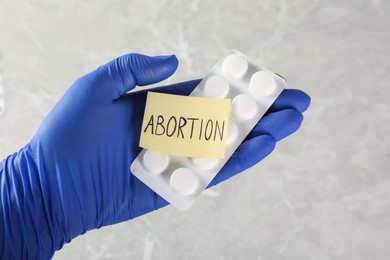 Photo of Doctor holding pills and paper card with word ABORTION on light grey background, closeup