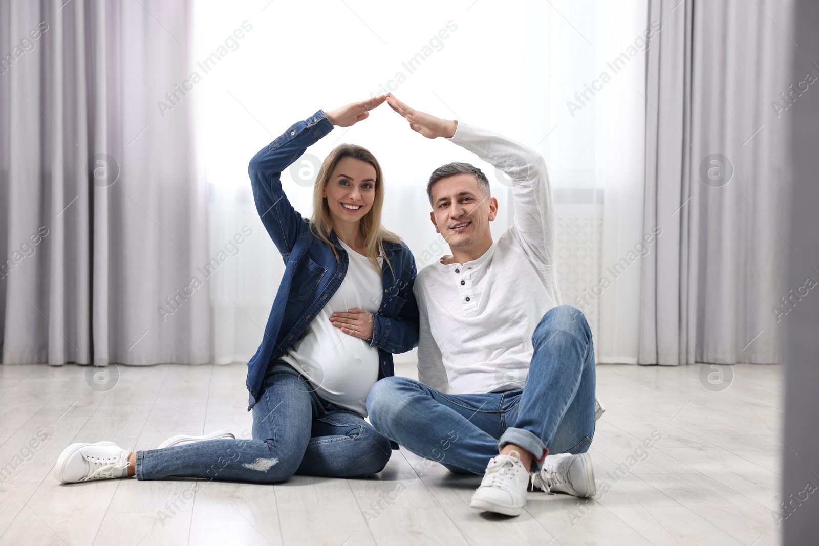 Photo of Young family housing concept. Pregnant woman with her husband forming roof with their hands while sitting on floor at home