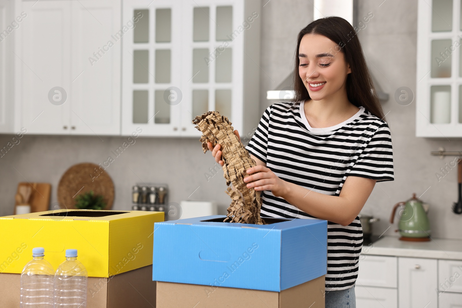 Photo of Garbage sorting. Smiling woman throwing shredded paper into cardboard box in kitchen