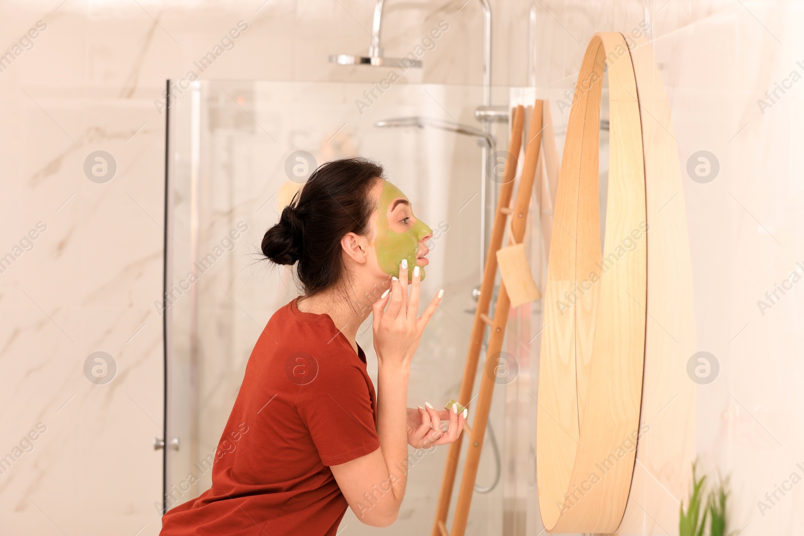 Photo of Young woman applying mask on her face near mirror in bathroom