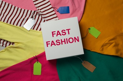 Photo of White box with phrase Fast Fashion and different clothes, top view