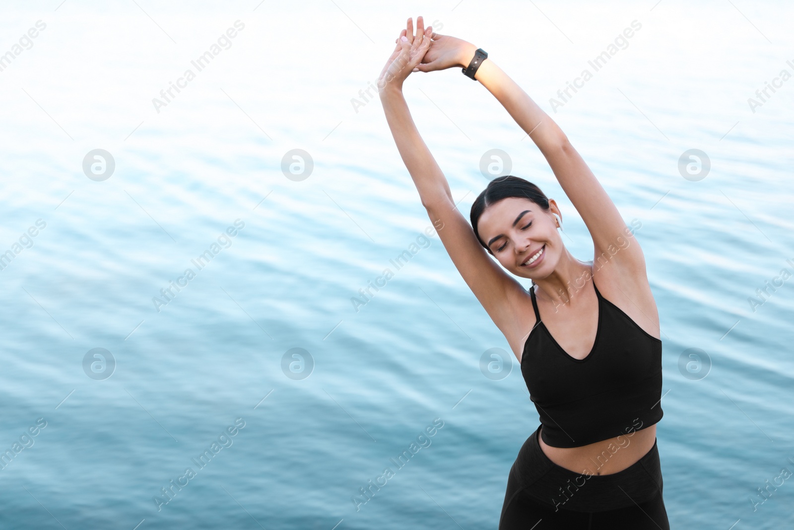 Photo of Young sportswoman with wireless earphones stretching near river