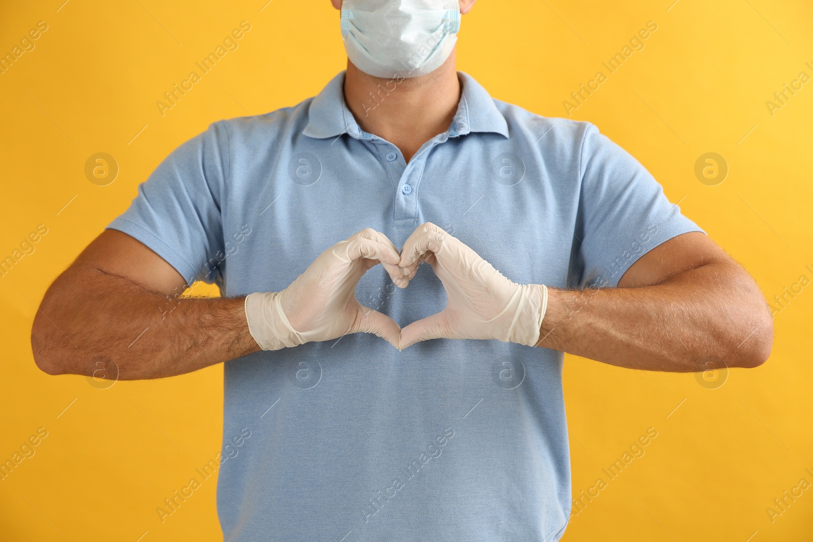 Photo of Man in protective face mask and medical gloves making heart with hands on yellow background, closeup