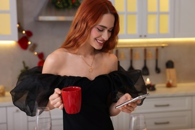 Beautiful young woman with cup of drink and smartphone in kitchen