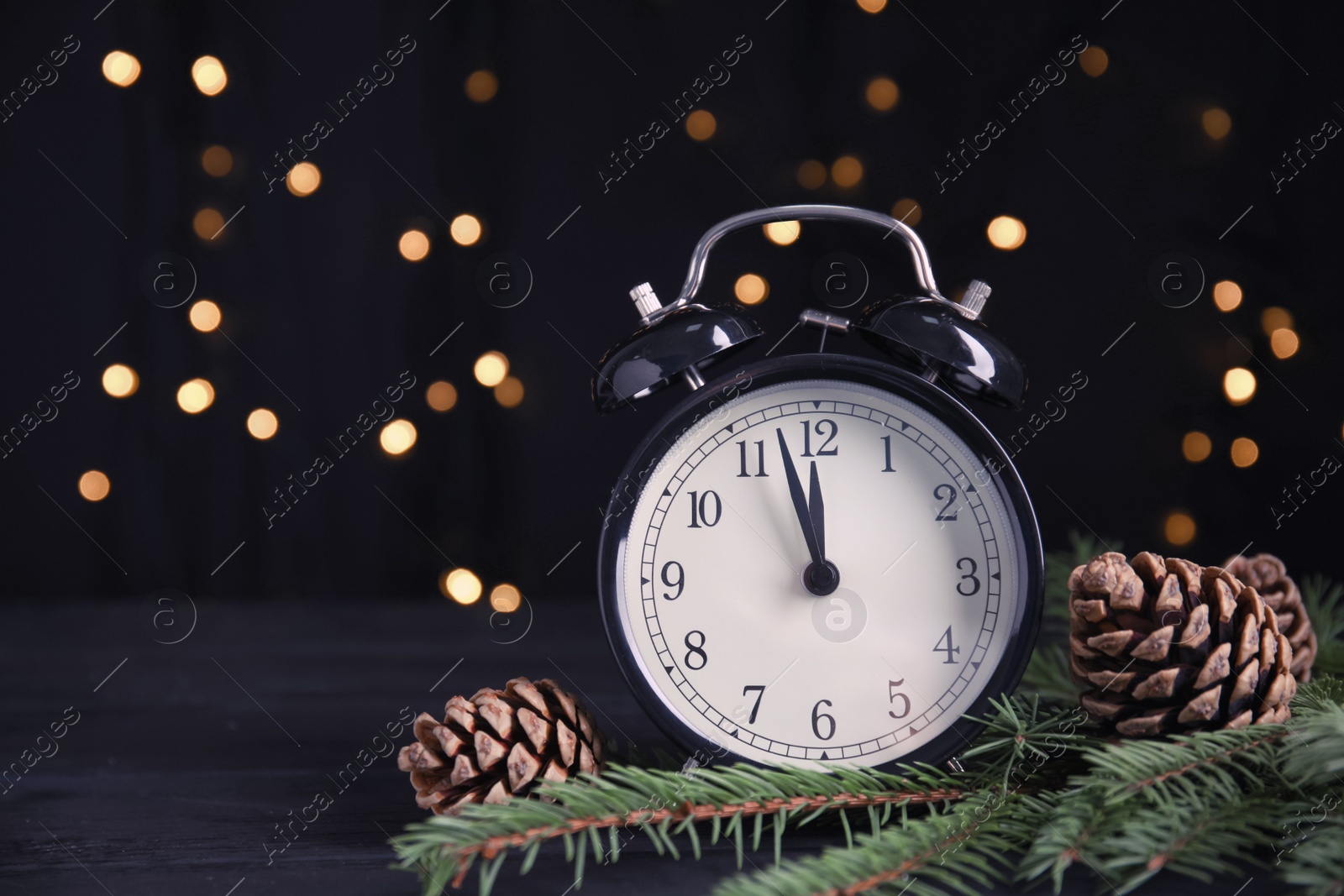 Photo of Christmas greeting card with space for text. Alarm clock, fir tree branches and pine cones on wooden table against blurred festive lights