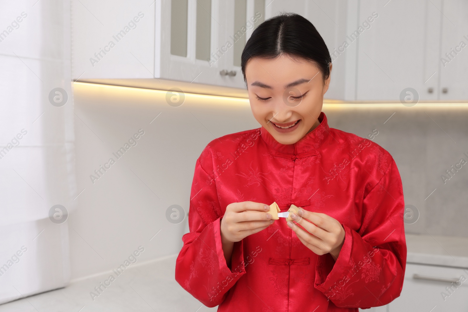 Photo of Asian woman holding tasty fortune cookie with prediction in kitchen. Space for text