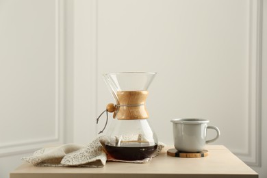Photo of Glass chemex coffeemaker with coffee and cup on table against white wall