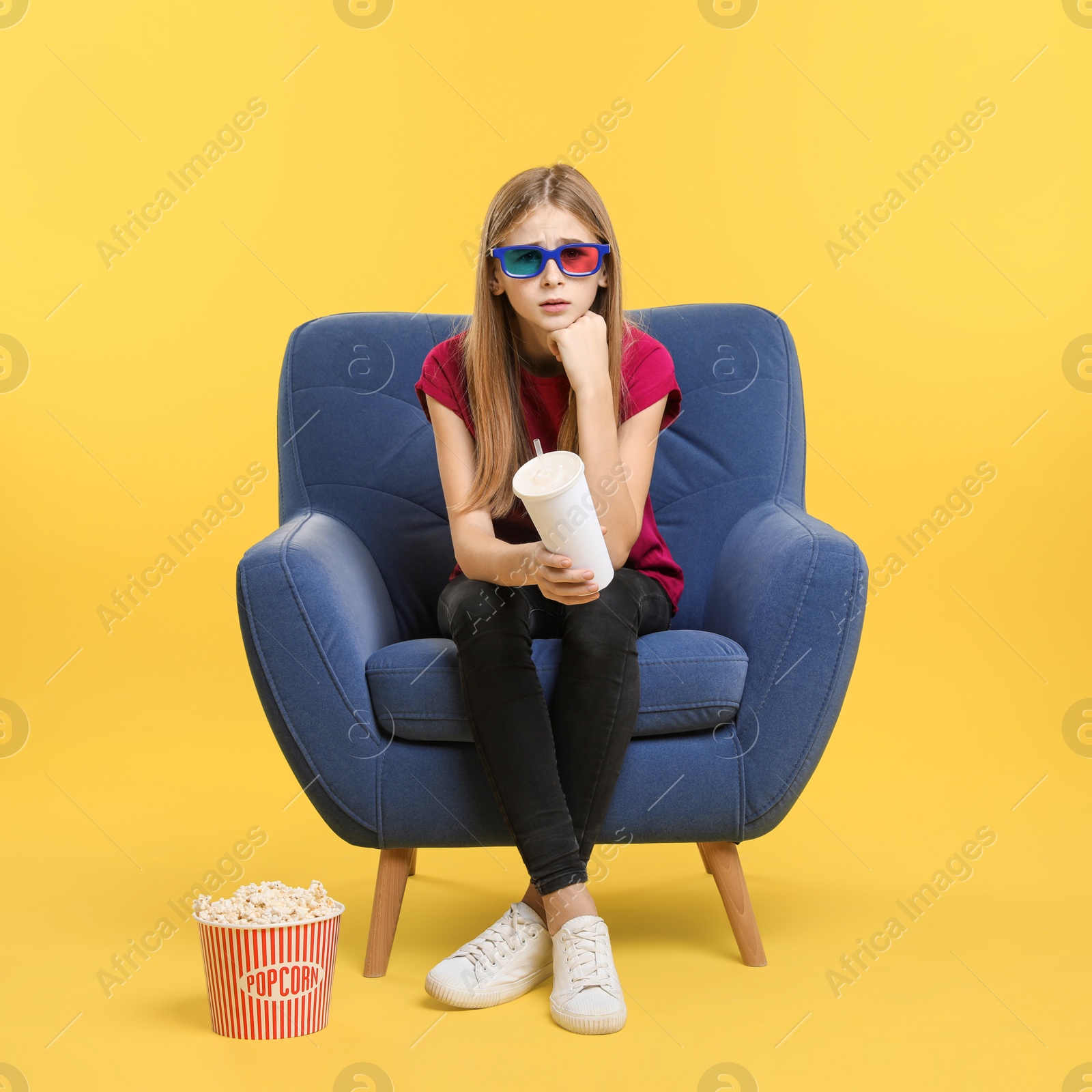 Photo of Emotional teenage girl with 3D glasses, popcorn and beverage sitting in armchair during cinema show on color background