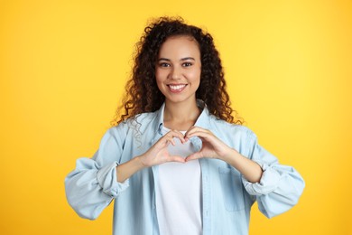 Photo of Happy young African-American woman making heart with hands on yellow background