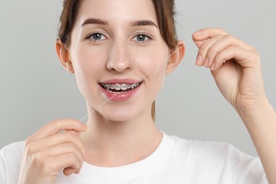 Smiling woman with braces cleaning teeth using dental floss on grey background, closeup