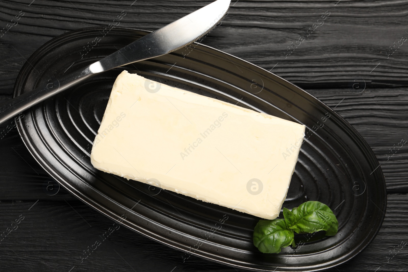 Photo of Block of tasty butter with basil and knife on black wooden table, top view