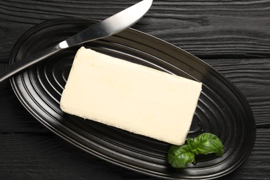 Block of tasty butter with basil and knife on black wooden table, top view