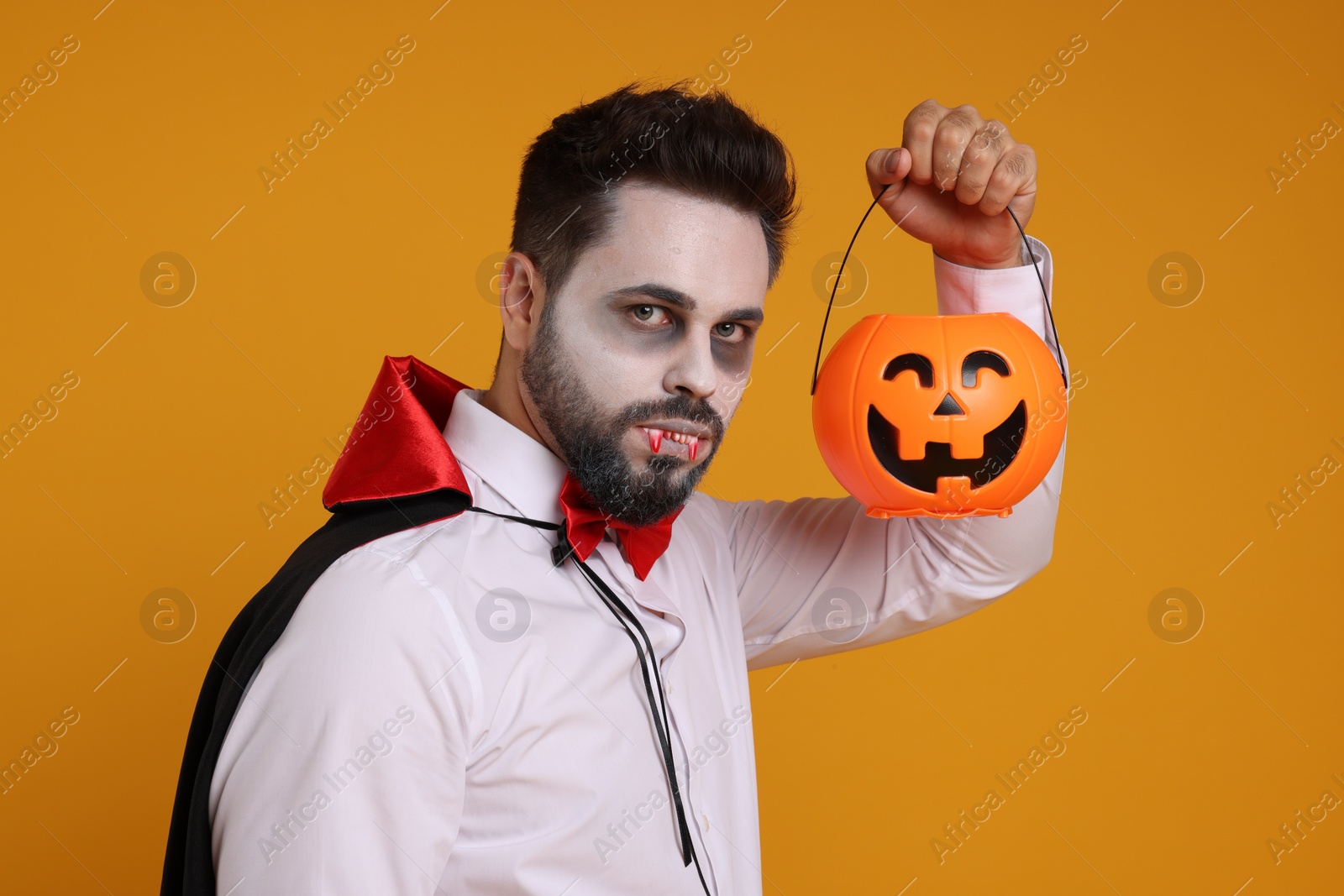 Photo of Man in scary vampire costume with fangs and pumpkin bucket on orange background. Halloween celebration