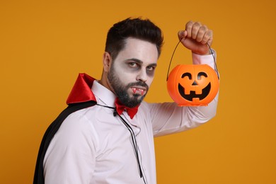 Photo of Man in scary vampire costume with fangs and pumpkin bucket on orange background. Halloween celebration