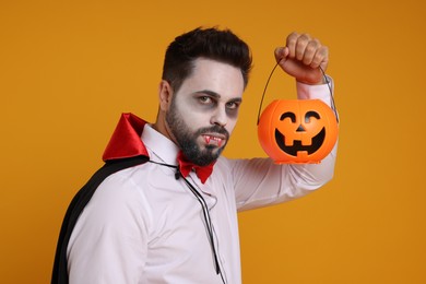 Man in scary vampire costume with fangs and pumpkin bucket on orange background. Halloween celebration