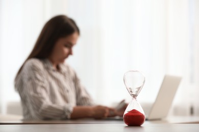 Photo of Hourglass with red flowing sand on table. Woman using laptop indoors, selective focus