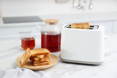 Photo of Modern toaster with slices of bread and tea on white marble table in kitchen