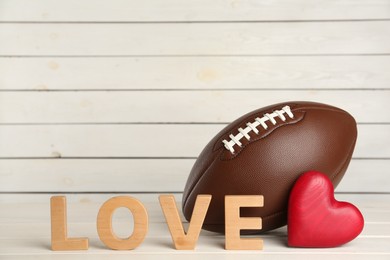 Photo of American football ball, heart and word Love on white wooden table. Space for text