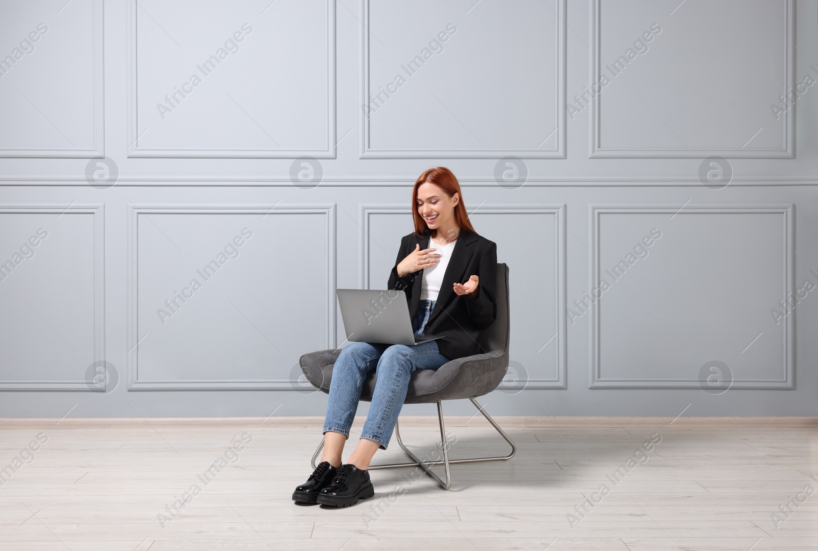 Photo of Happy young woman having video chat via laptop while sitting in armchair near light grey wall indoors