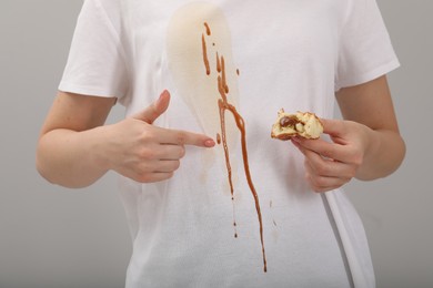 Woman holding pastry and showing stain from condensed milk on her shirt against light grey background, closeup