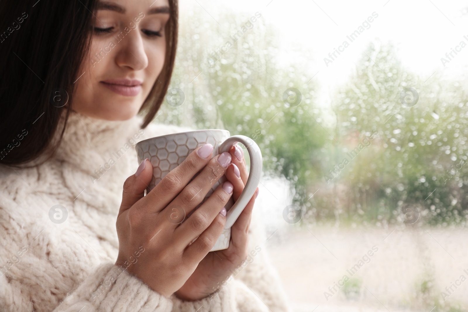 Photo of Beautiful woman with cup of tea near window indoors on rainy day, closeup