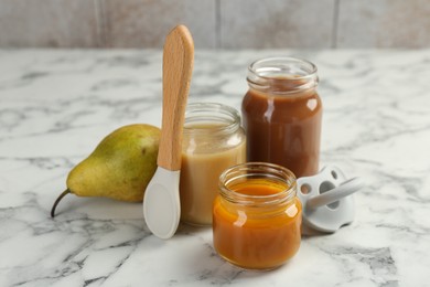 Tasty baby food in jars, pear, pacifier and spoon on white marble table