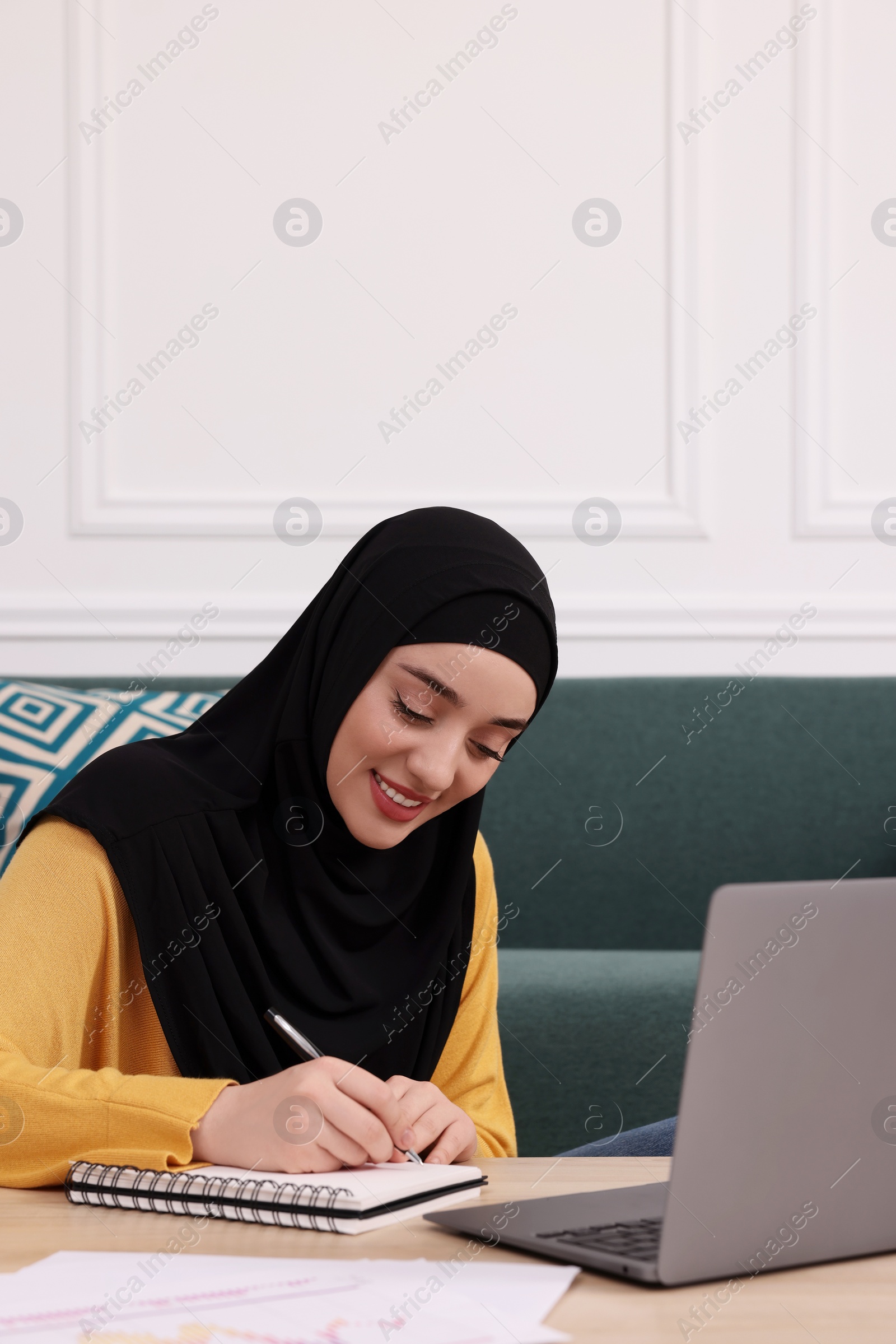 Photo of Muslim woman in hijab writing notes near laptop at wooden table indoors