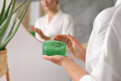 Photo of Young woman holding jar of aloe gel near mirror in bathroom, closeup