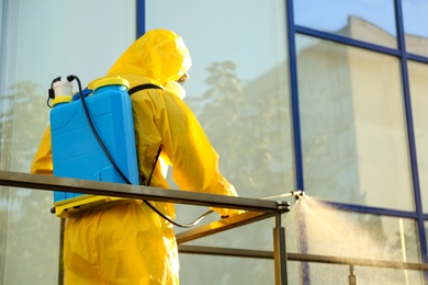 Person in hazmat suit with disinfectant sprayer cleaning metal railing on city street. Surface treatment during coronavirus pandemic