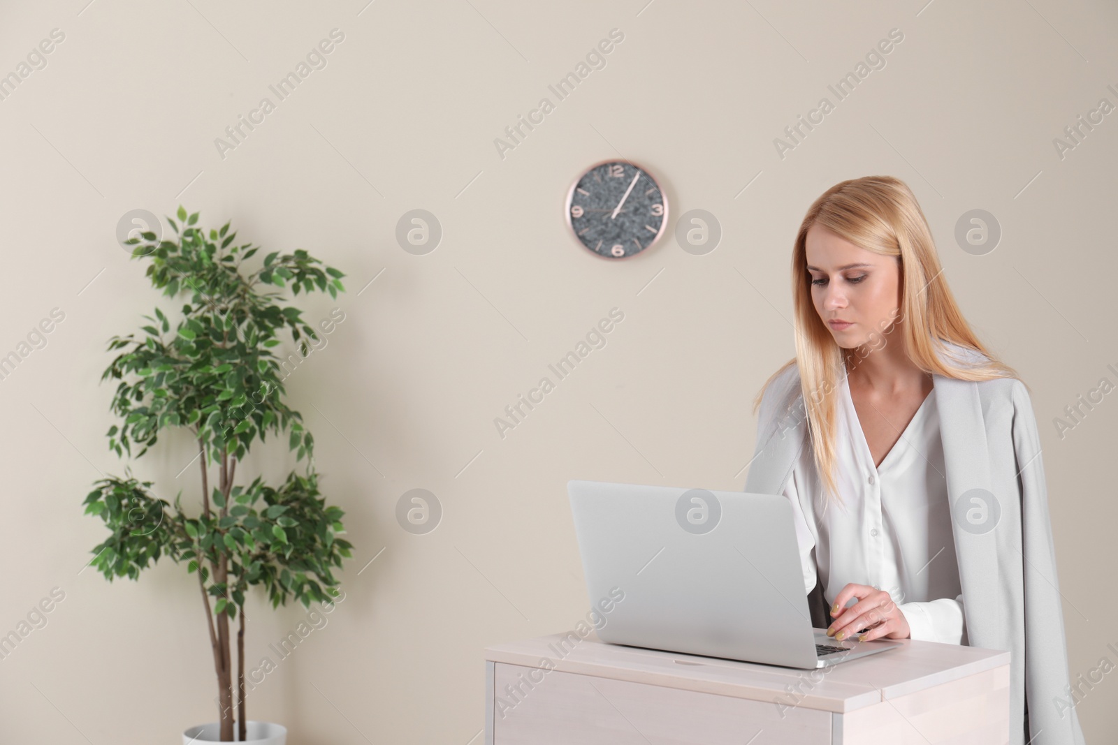 Photo of Young woman using laptop at stand up workplace against light wall
