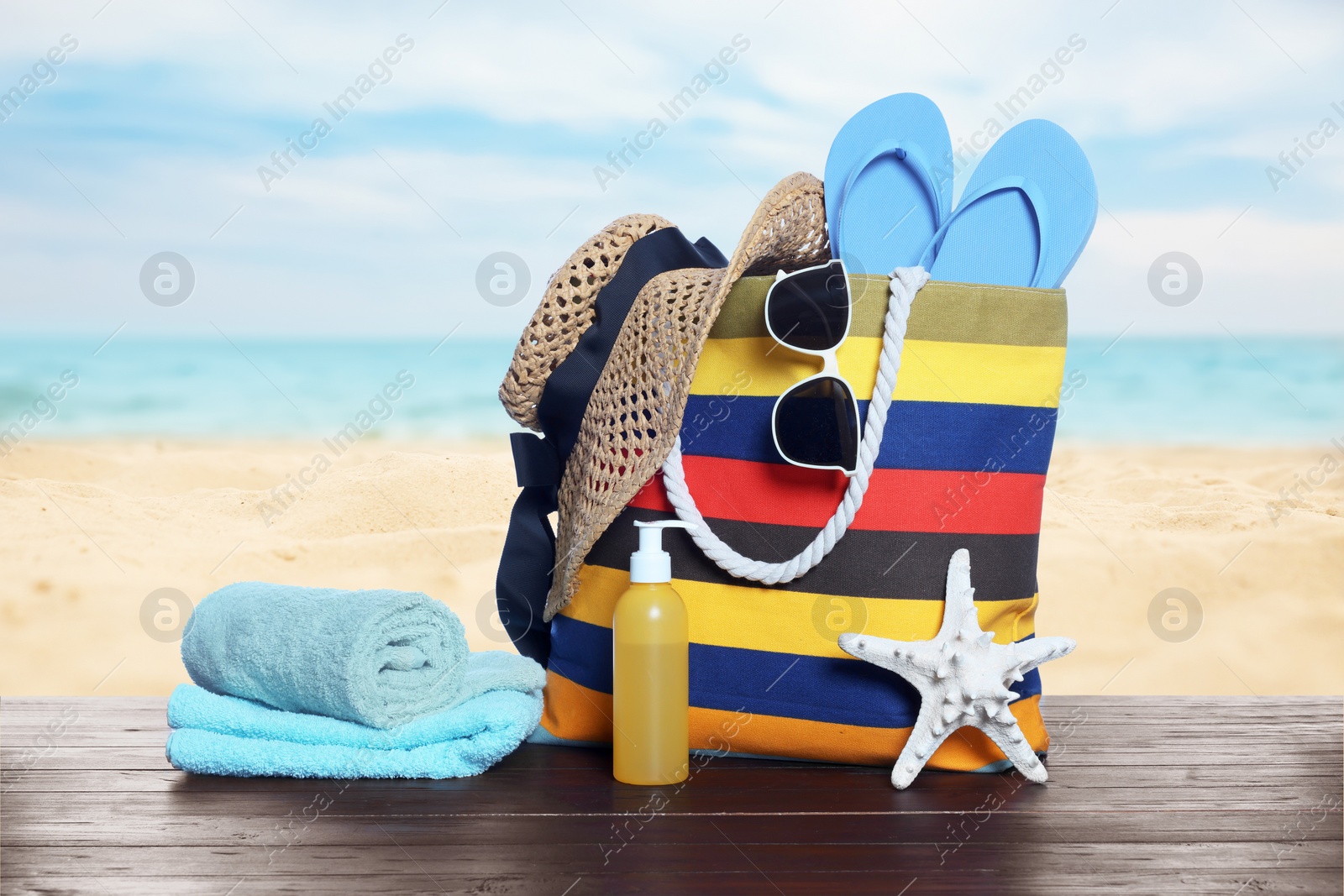 Image of Stylish striped bag and other beach accessories on wooden table near sea