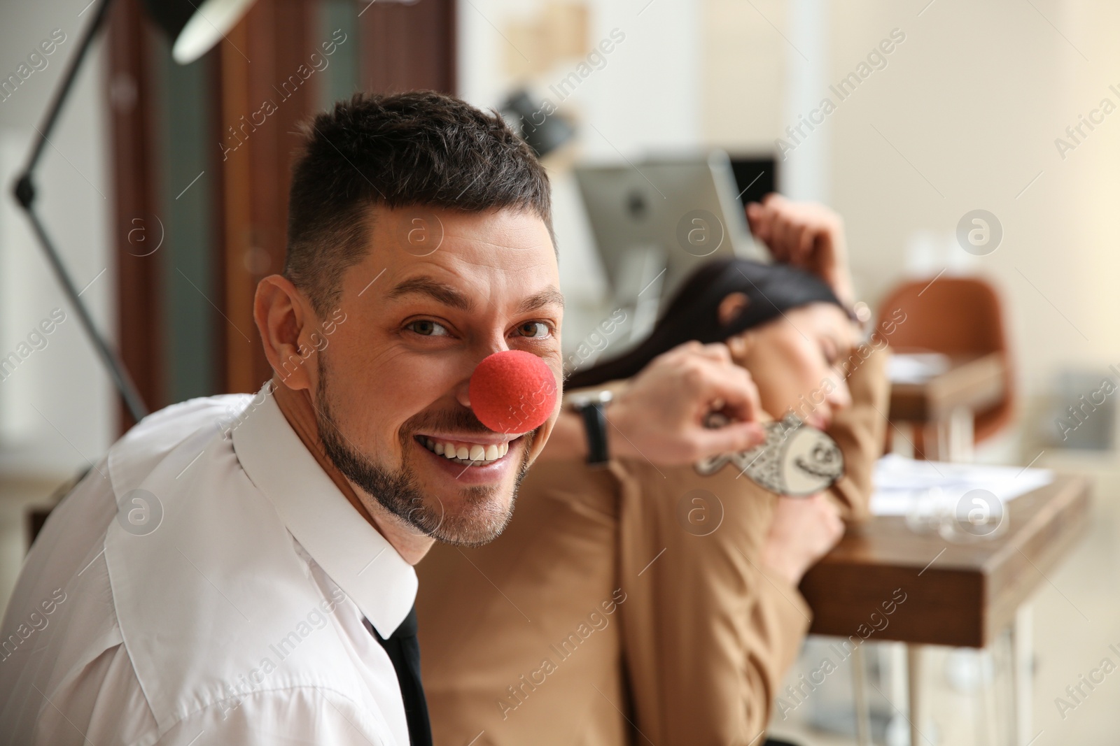 Photo of Man with clown nose sticking paper fish to colleague's back while she sleeping in office. Funny joke