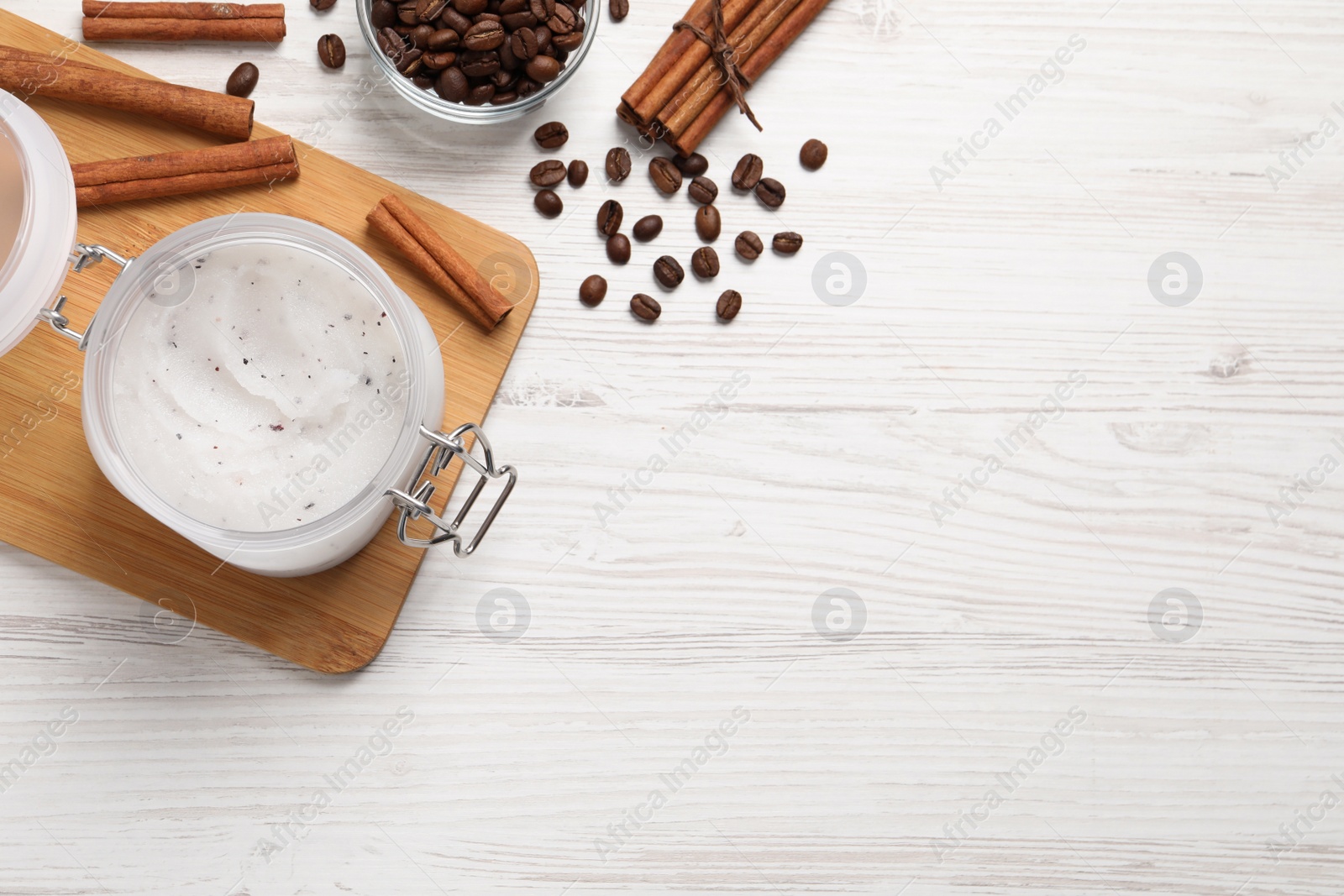 Photo of Body scrub in glass jar, cinnamon and coffee beans on white wooden table, flat lay. Space for text