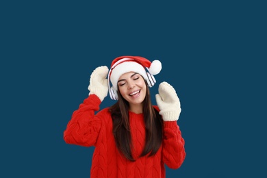 Young woman in Santa hat listening to Christmas music on dark blue background