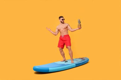 Photo of Happy man with pineapple posing on SUP board against orange background