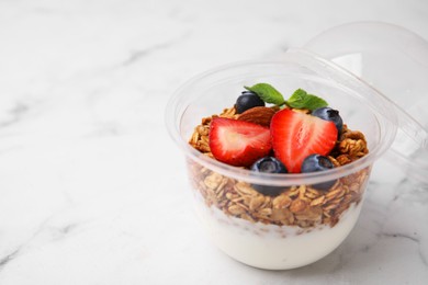 Photo of Tasty granola with berries and yogurt in plastic cup on white marble table, closeup. Space for text