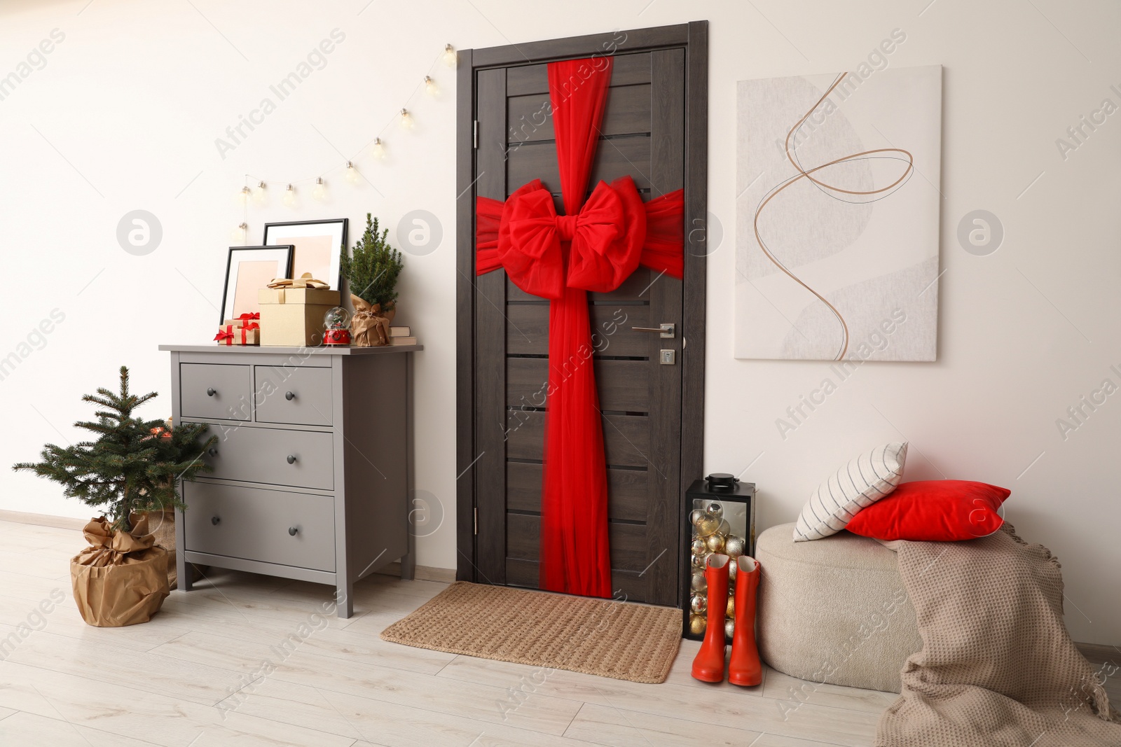 Photo of Wooden door with beautiful red bow near chest of drawers, evergreen trees and gift boxes indoors. Christmas decoration